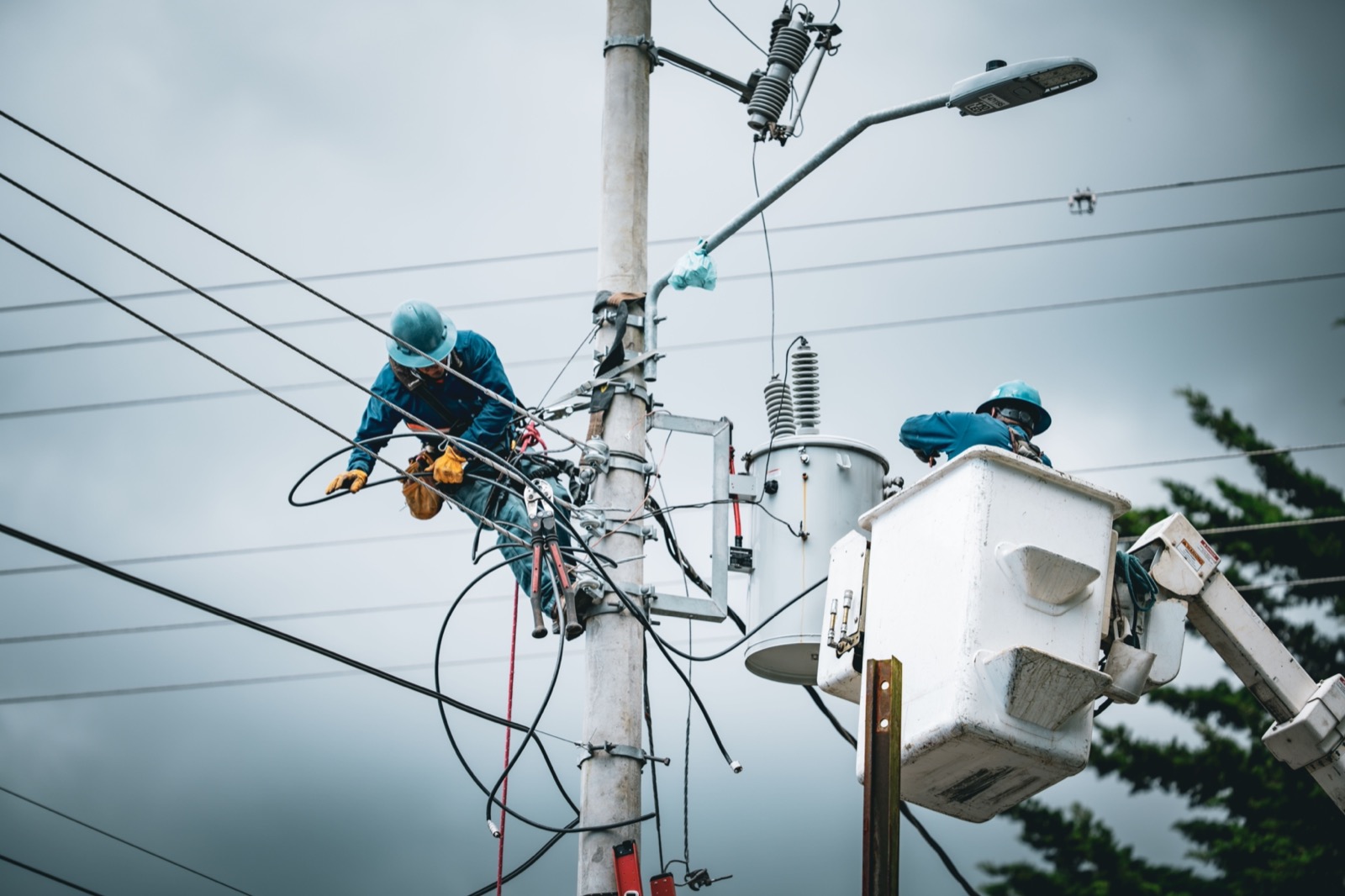 Two linemen in blue hard hats working on a concrete utility pole beside a pole mounted transformer, one on the pole and one in a bucket lift
