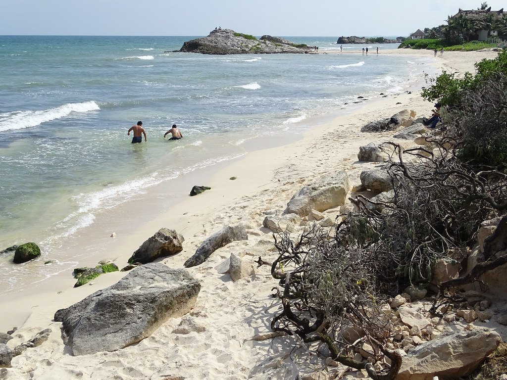 Two swimmers in clear shallow water off a wide, clean stretch of Playa Tulum, with limestone rocks and low scrub along the sand