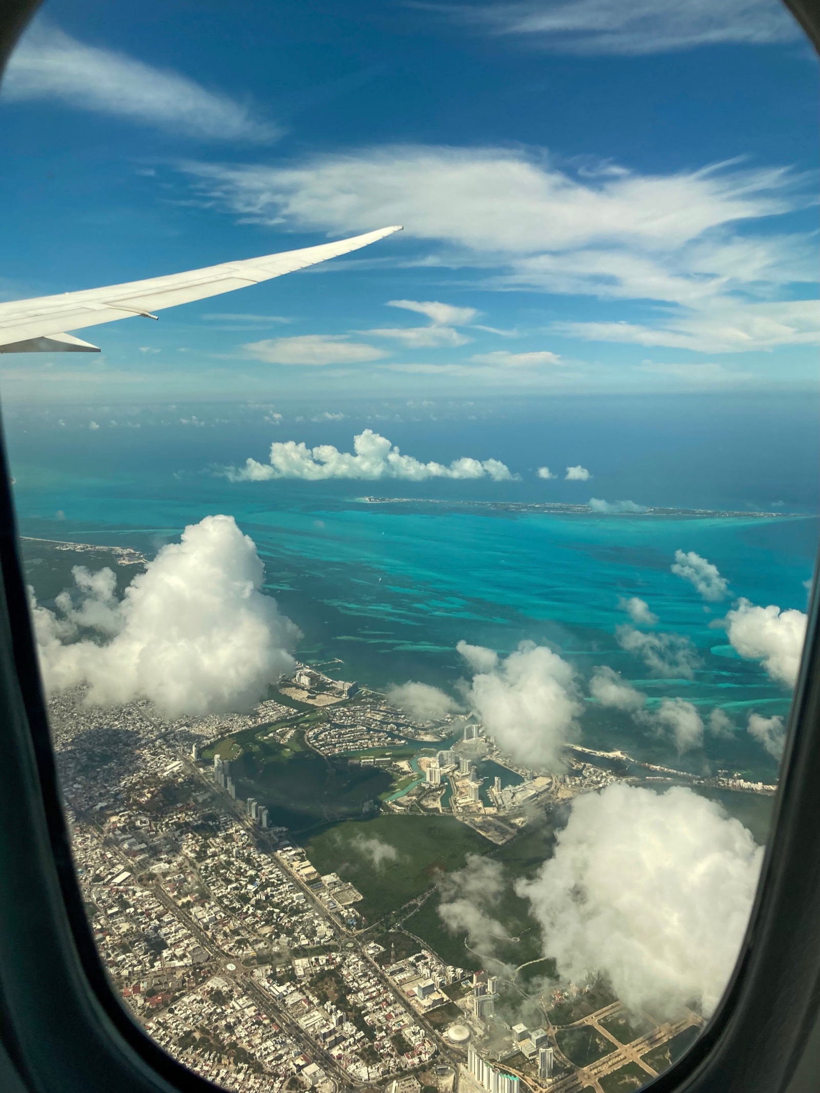 View through an aircraft window on approach to Cancún, with the wing at upper left over turquoise shallows, the Puerto Cancún marina district and Isla Mujeres on the horizon