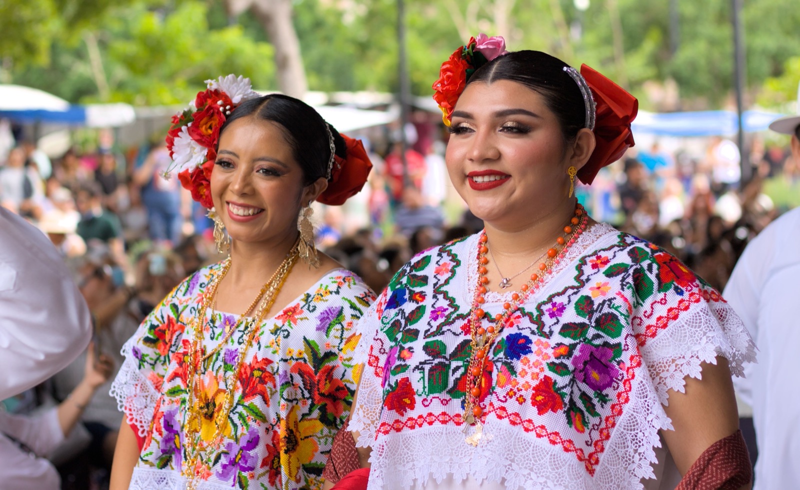 Two women in white Yucatecan terno dresses with cross-stitch floral embroidery, red flowers in their hair and coral necklaces, smiling in front of a blurred festival crowd under trees
