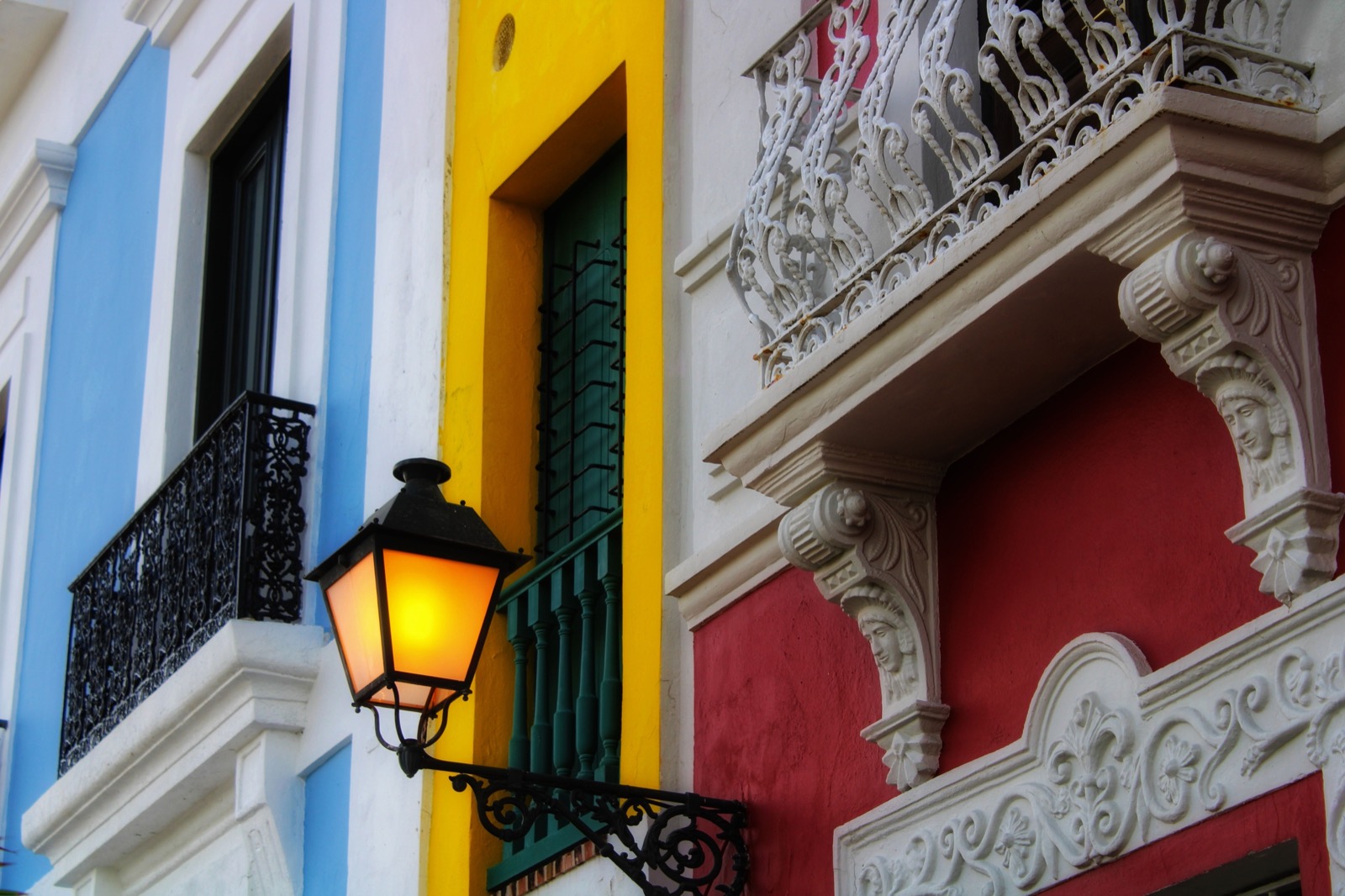 Close view of brightly painted Caribbean-style house facades in blue, yellow and red, with wrought-iron balconies, green shuttered windows and a lit wall lantern
