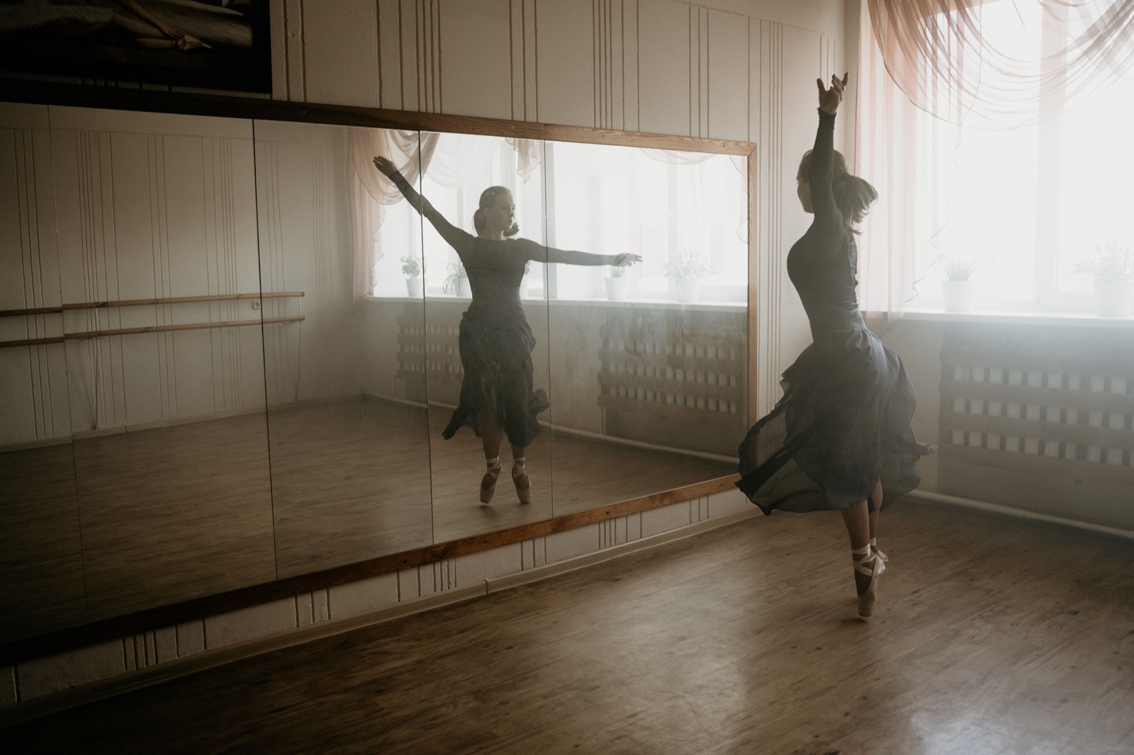 A dancer in a dark dress rehearsing alone in a mirrored teaching studio with a ballet barre and a worn wooden floor, her movement doubled in the glass