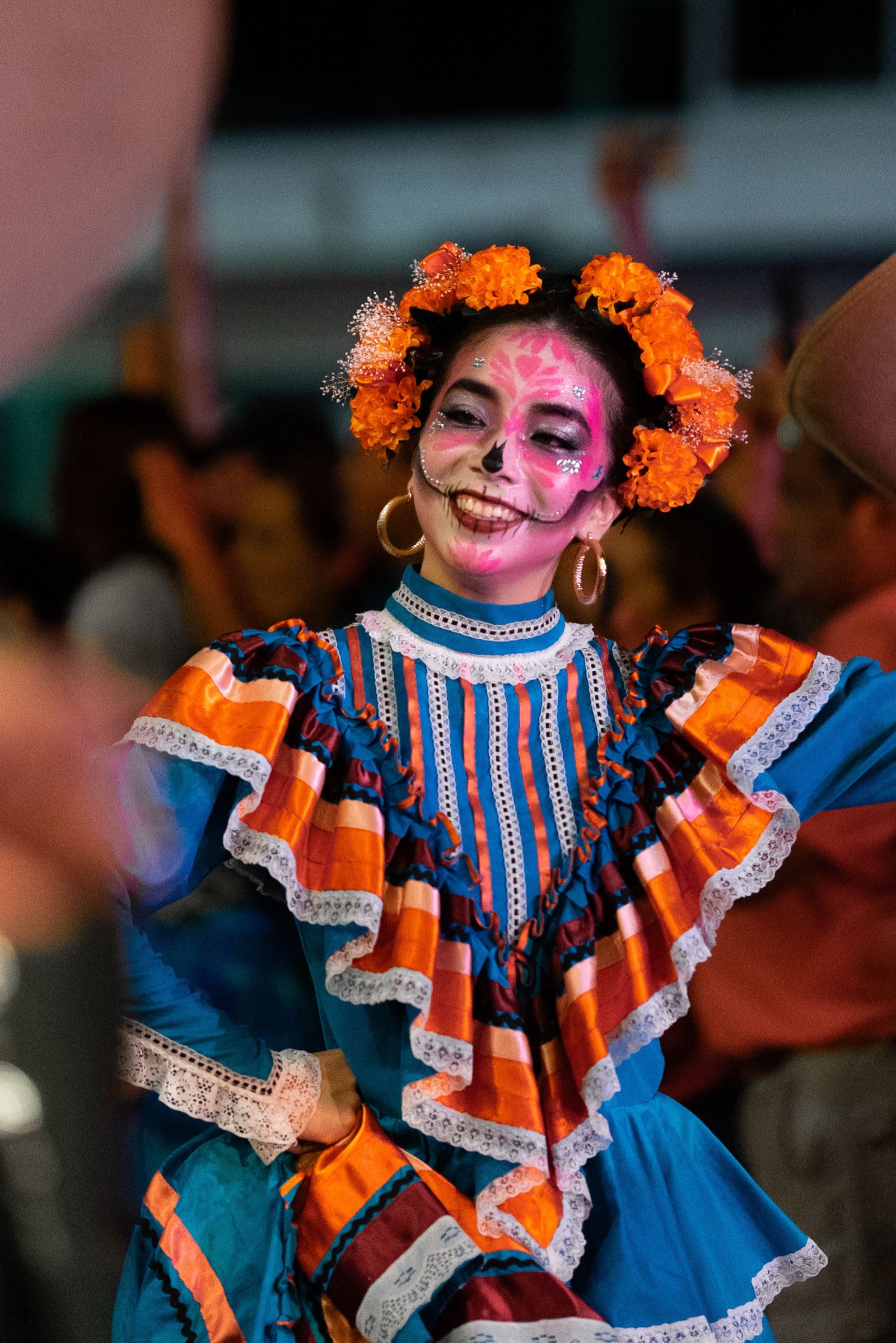 A young woman at night wearing calavera face paint, an orange marigold crown and a blue folkloric dress, with a blurred crowd behind her