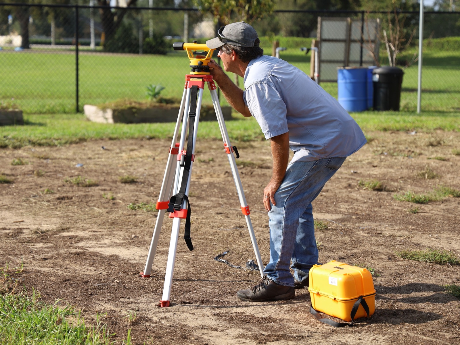A man in a cap and jeans working at a yellow surveyor's level on a tripod, on a bare dirt plot with a chain-link fence and trees behind