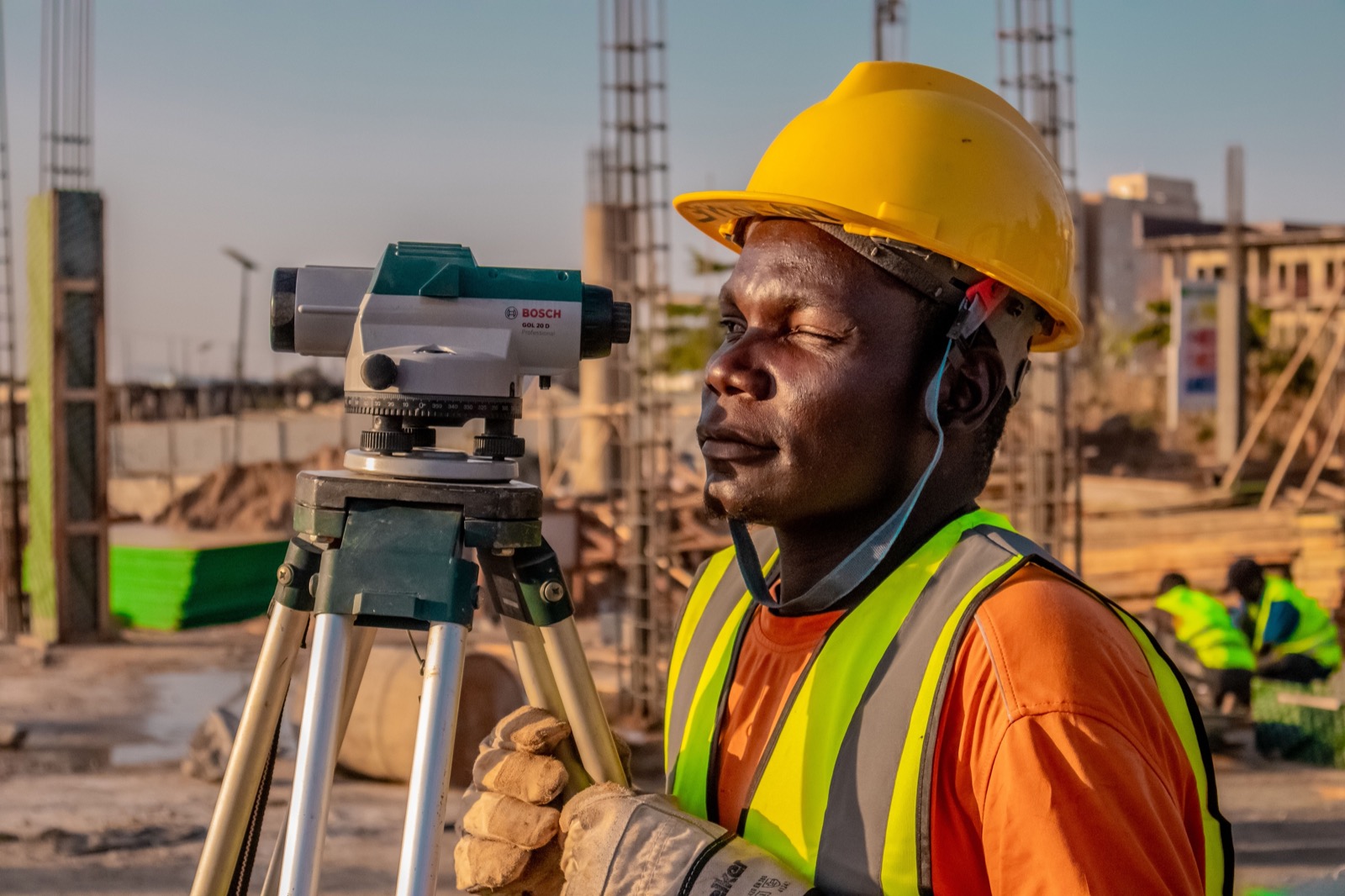 A surveyor in a yellow hard hat and high-visibility vest stands beside an optical level on a tripod on a construction site
