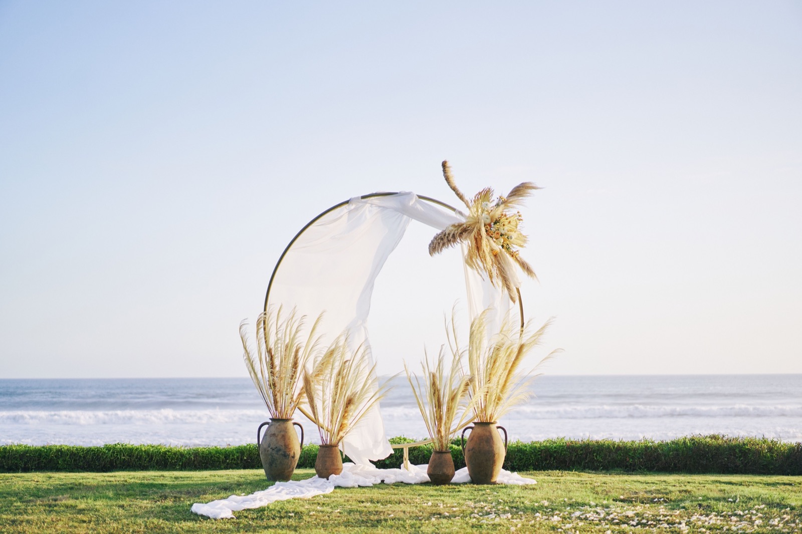 A ceremony arch draped in white fabric and pampas grass, set on a lawn above the ocean for a seaside wedding