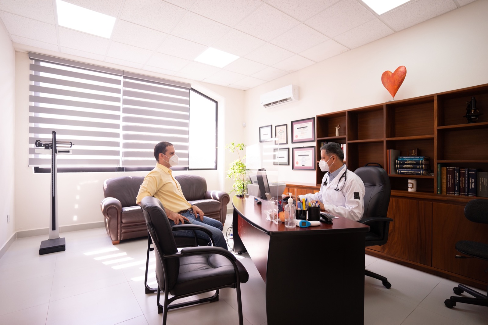 A doctor in a white coat and face mask consults with a patient across a desk in a bright clinic room