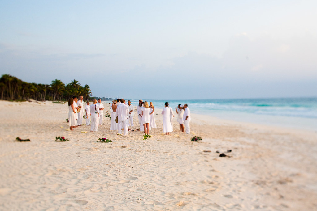 Wedding guests dressed in white gather in a loose circle on a wide sandy beach at dawn, with flowers laid on the sand and palms along the shore