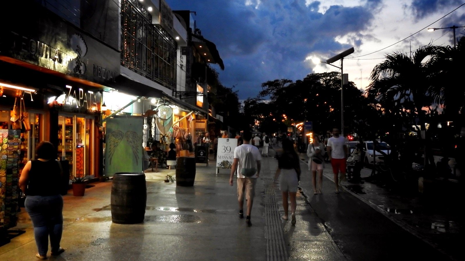 Shops lit up at dusk along a wet sidewalk on Avenida Tulum, with people walking under a cloudy evening sky