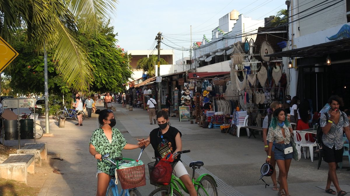 Shops with hammocks, hats and textiles along a wide sidewalk on Avenida Tulum, with people walking and two women on bicycles