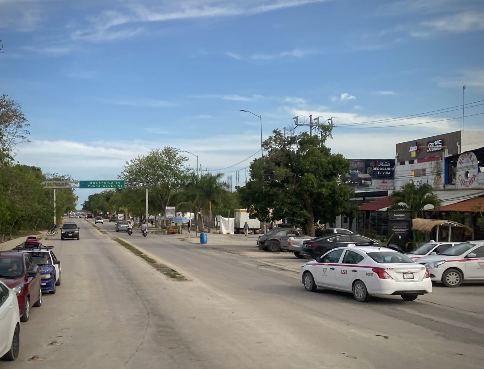 A wide road at the edge of Tulum town with taxis parked on the right and a green overhead sign reading Bocapaila 20, Punta Allen 56