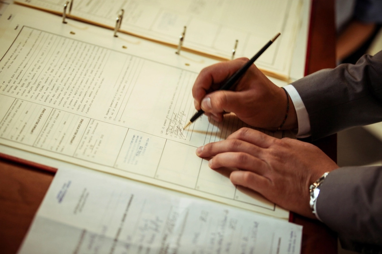 A hand signing a contract in a bound register, with loose documents beside it