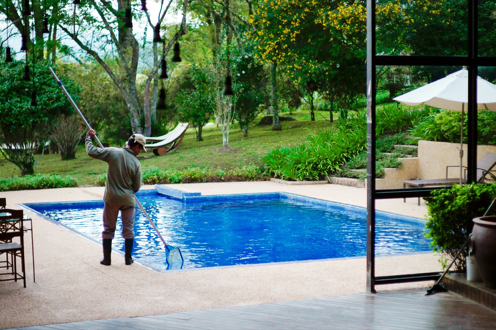 A worker in a cap and rubber boots skimming a small outdoor swimming pool with a long-handled net, beside a lawn, sun loungers and a patio umbrella.