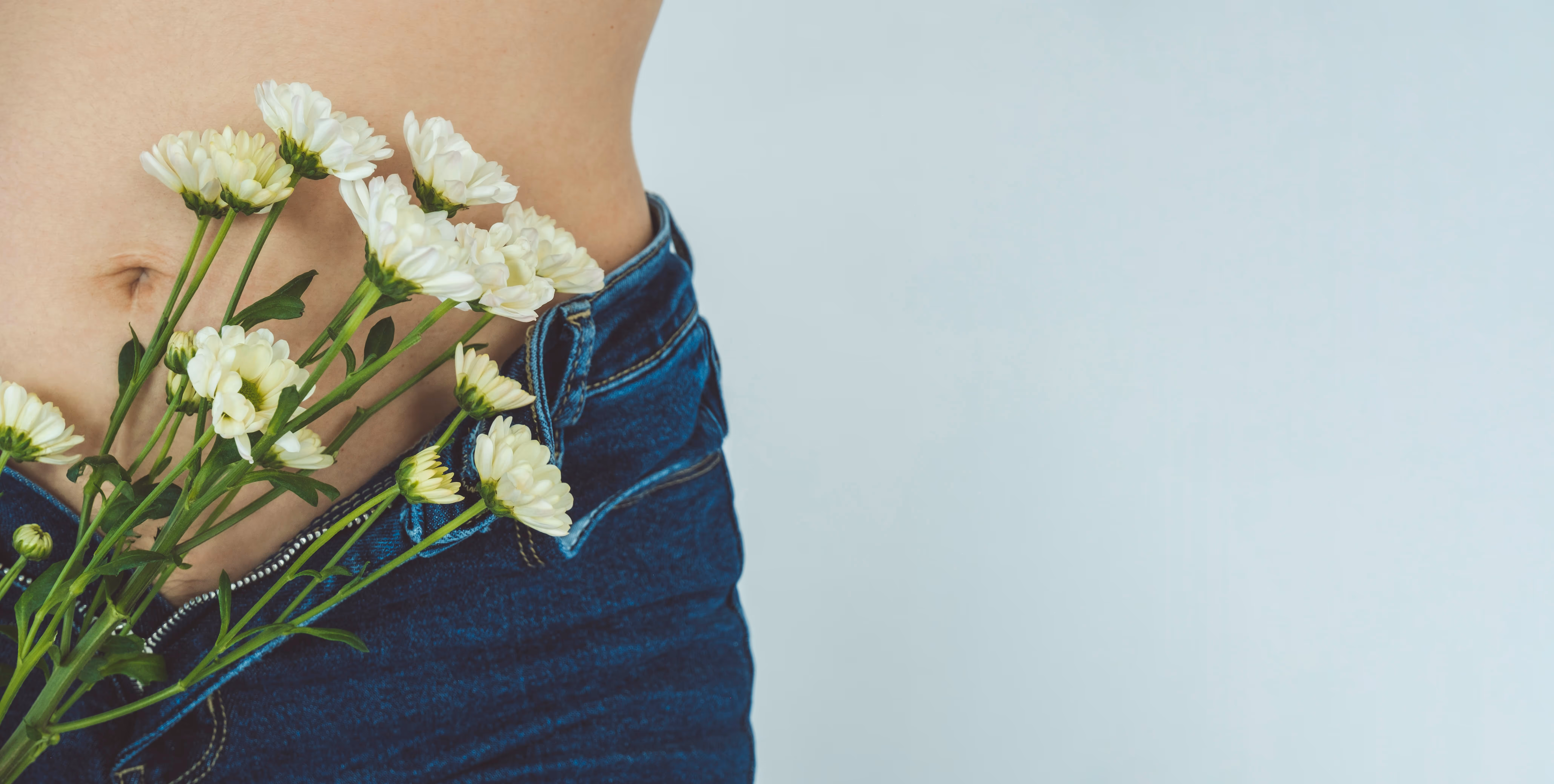 A close-up of a person's midsection with a bouquet of white daisies tucked into the waistband of blue jeans against a light blue background.