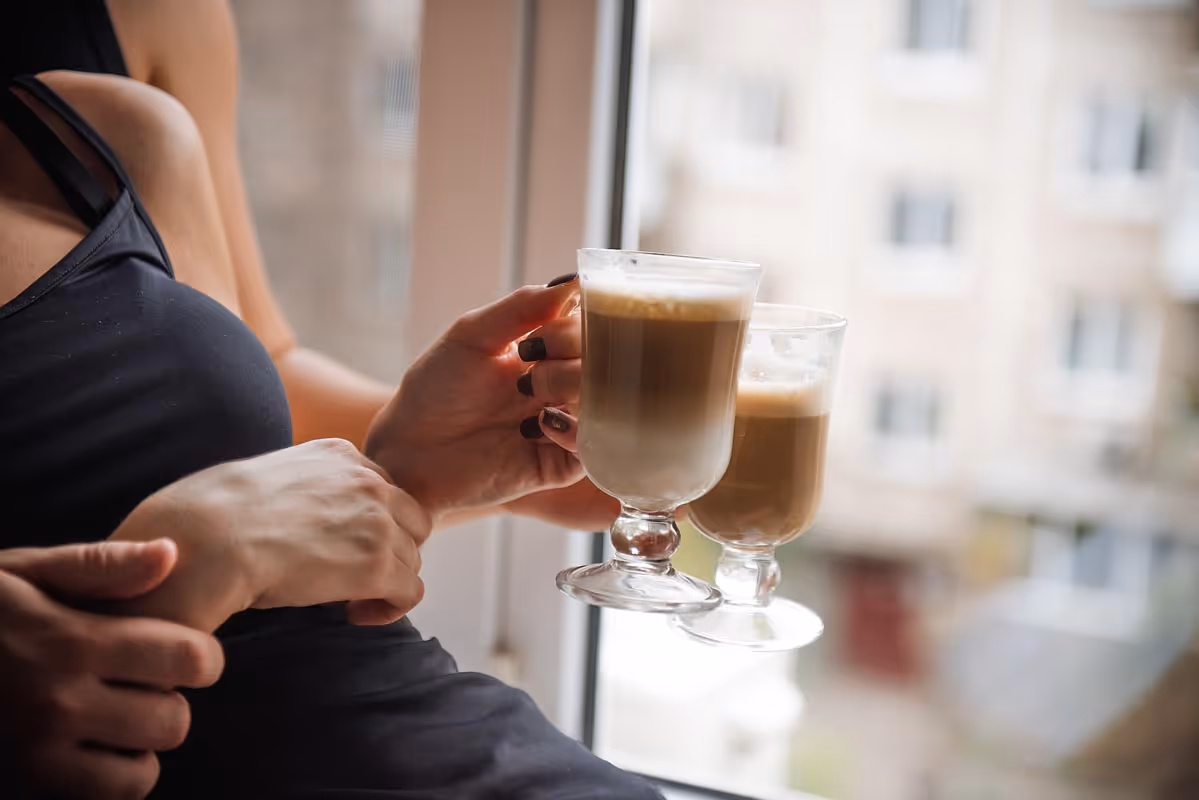 A person in a black top holding two glasses of iced coffee, with another hand resting on their arm, against a window with a blurred view of buildings outside.