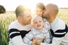 Two men in white shirts with black stripes hugging two young children in a field of tall grass, with the sun shining brightly in the background.
