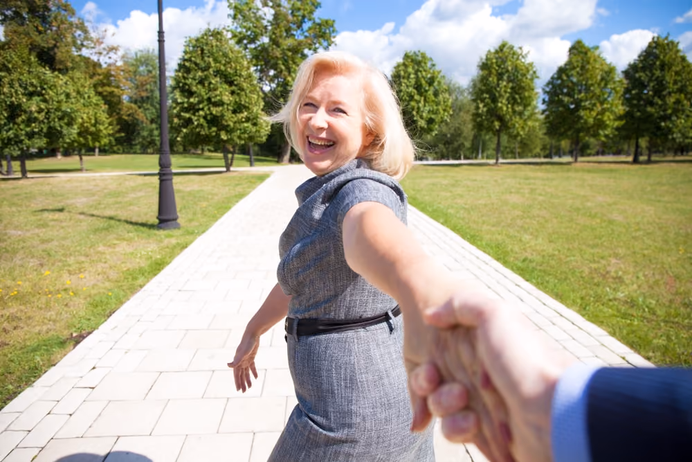 A smiling woman in a gray dress holds someone's hand while walking on a paved path in a park with green trees and a blue sky.