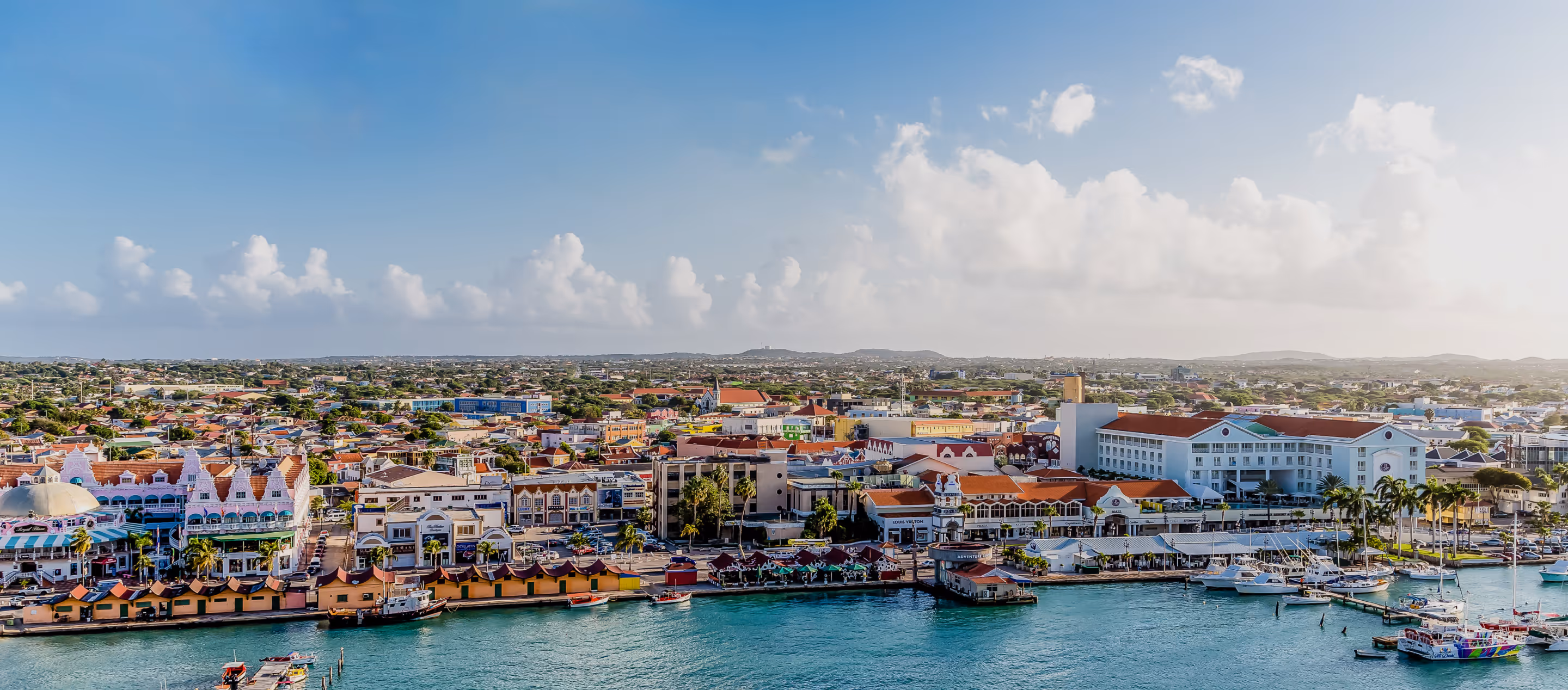 Panoramic view of Oranjestad, Aruba, with colorful waterfront buildings, marina, and turquoise harbor under a clear blue sky.