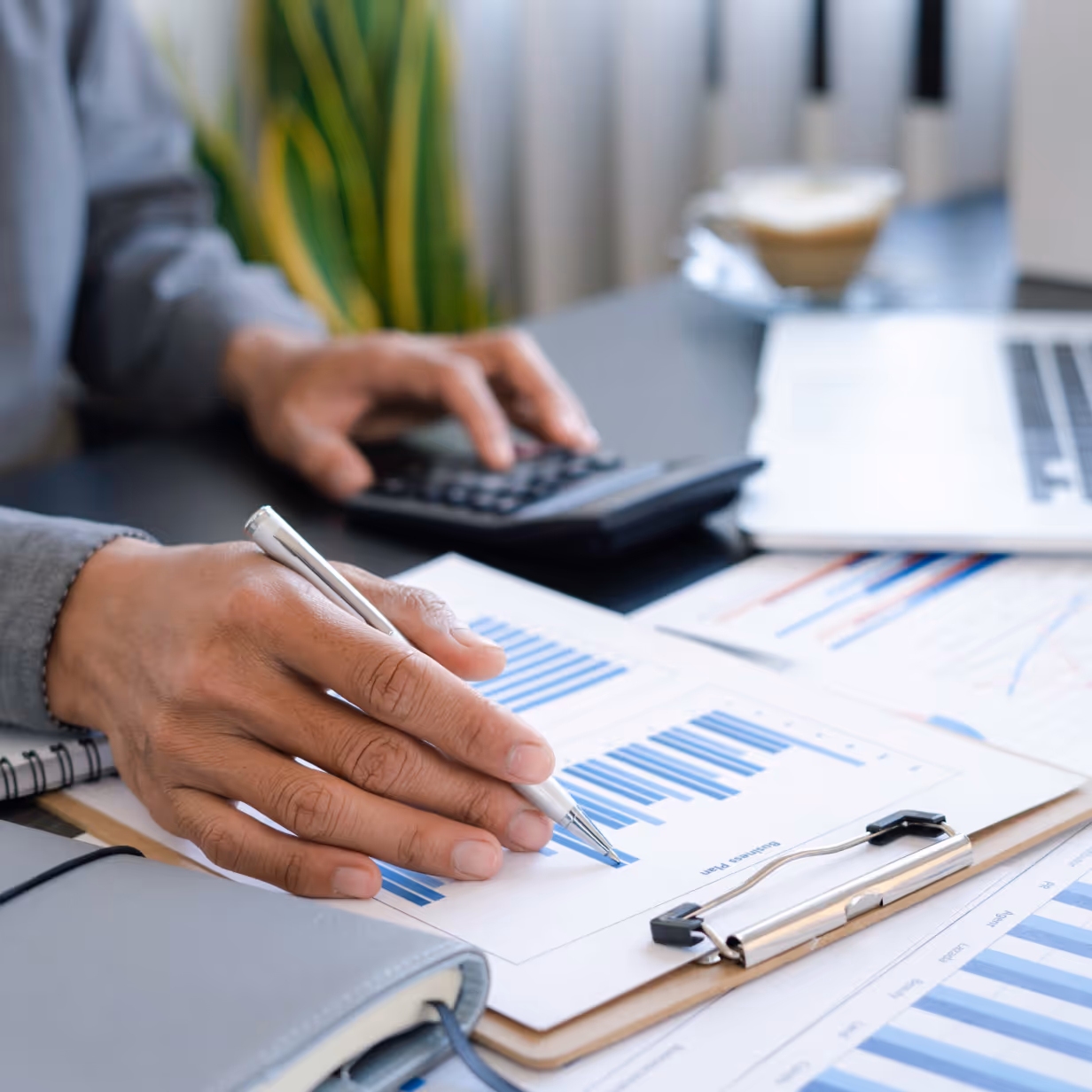 Close-up of hands reviewing financial charts and graphs on a clipboard, with a calculator and laptop on the desk, representing careful analysis and strategic decision-making.