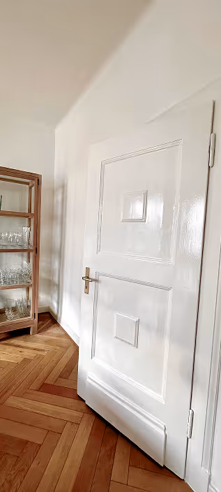 Weiße Zimmertür mit goldfarbenem Griff neben einem Holzschrank mit Glasgeschirr auf einem Parkettboden.