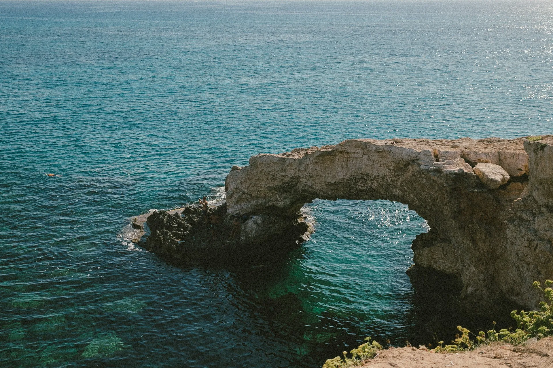 Natural stone arch over clear blue sea with rocky cliffs and a swimmer in the water.