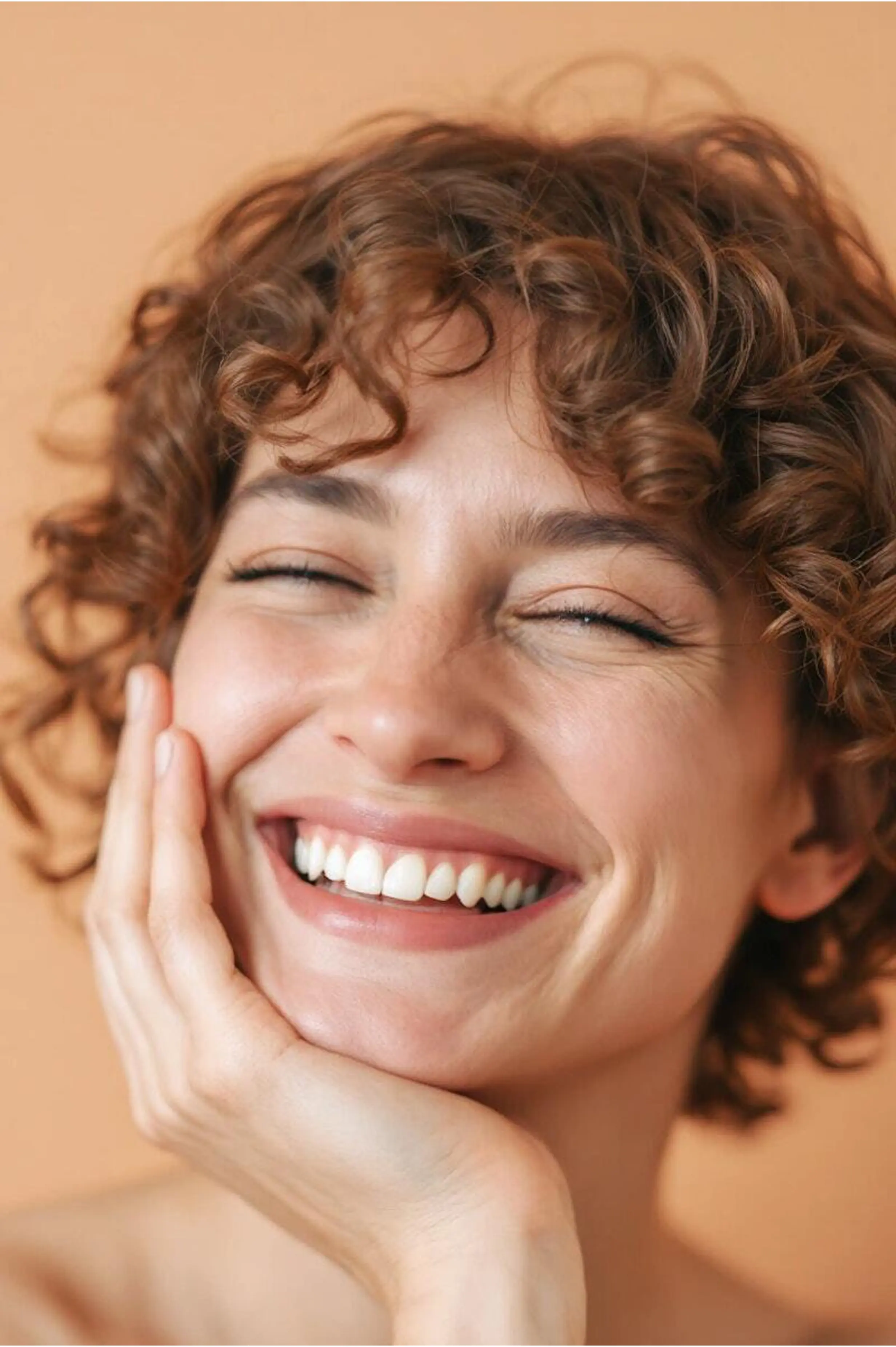 A person with short, curly hair smiles broadly with their eyes closed, teeth showing, resting their hand on their cheek against a plain background.