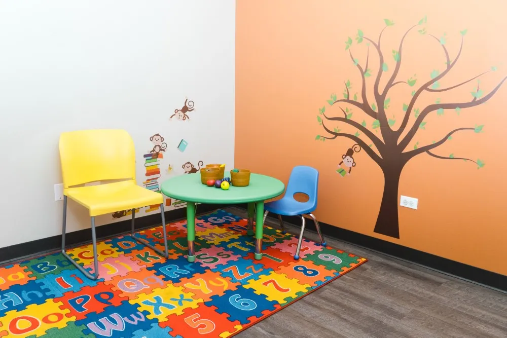 Children's playroom corner with colorful decor, a yellow and a blue chair, a round green table, a tree wall decal, and an alphabet mat.