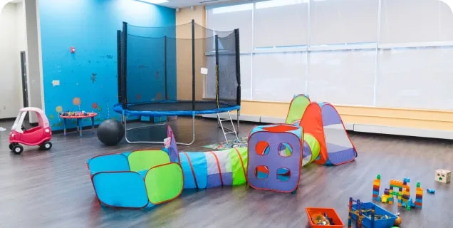 Brightly colored indoor playroom with a trampoline, play tunnel set, toy car, and scattered toys.