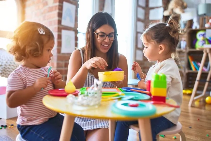 Woman and two children playing with toys at a small yellow table in a well-lit room.