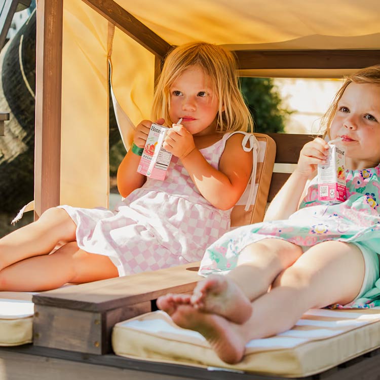 Two young girls relaxing and sipping juice boxes under a shaded play structure