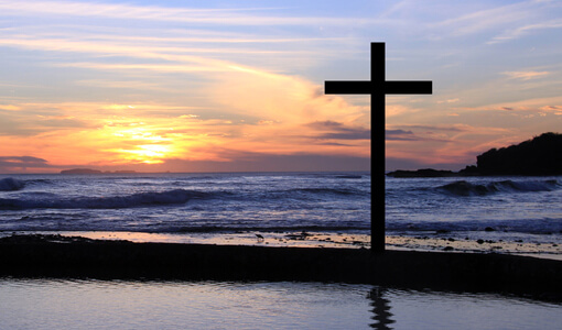 A cross in silhouette with the sun setting over gentle ocean waves