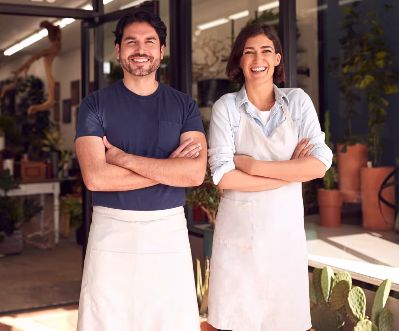 Homem e mulher sorridentes usando aventais, lado a lado com os braços cruzados na frente de uma loja com plantas.