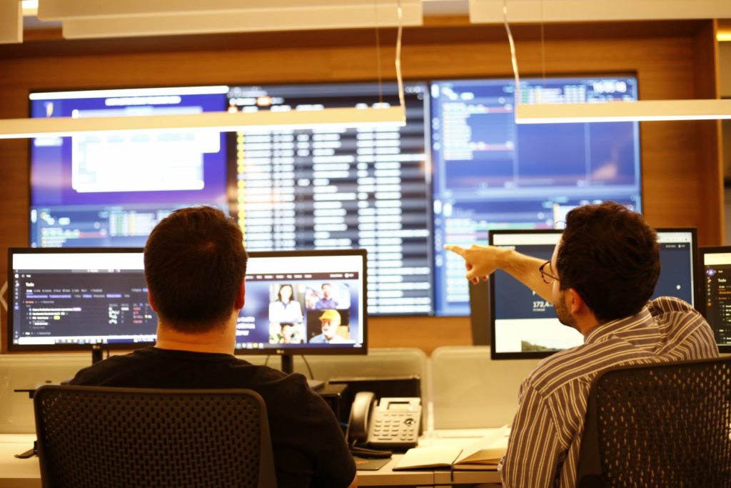 Duas pessoas em uma sala de controle observando múltiplos monitores com dados e gráficos, uma delas aponta para uma tela grande ao fundo.