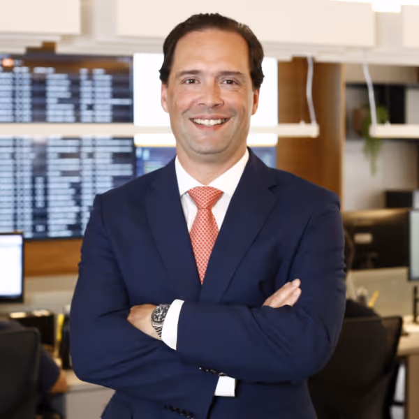 Homem sorridente de terno azul e gravata laranja com os braços cruzados em escritório moderno.