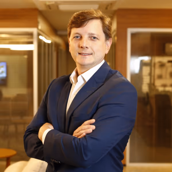 Homem vestido com terno azul e camisa branca, braços cruzados, sorrindo em escritório moderno.