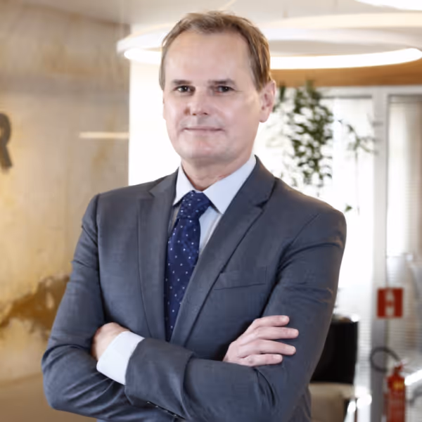 Homem de terno cinza com gravata azul escura posando com os braços cruzados em um ambiente de escritório moderno.