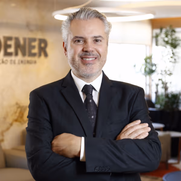 Homem de meia-idade com cabelo grisalho, vestindo terno preto e gravata, sorrindo com os braços cruzados em escritório moderno.