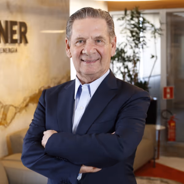 Homem de terno azul com os braços cruzados sorrindo em um escritório moderno.