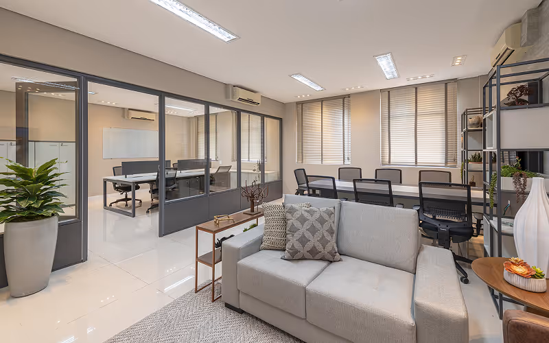 Sala de escritório moderna com sofá cinza, mesa de reunião, cadeiras giratórias pretas e grande janela com persianas.