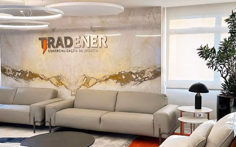 Sala de espera moderna com três sofás cinza, mesa de centro preta, lâmpada preta e planta ao lado da janela.