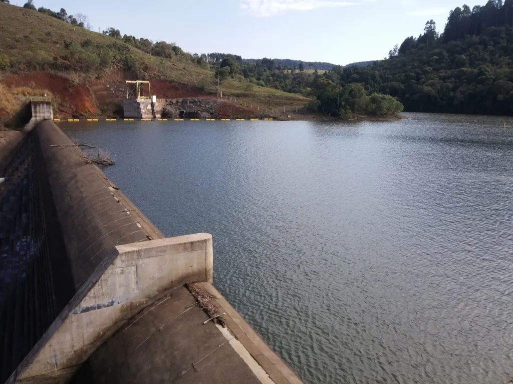Represa de concreto com água azul e colinas verdes ao redor sob céu claro.