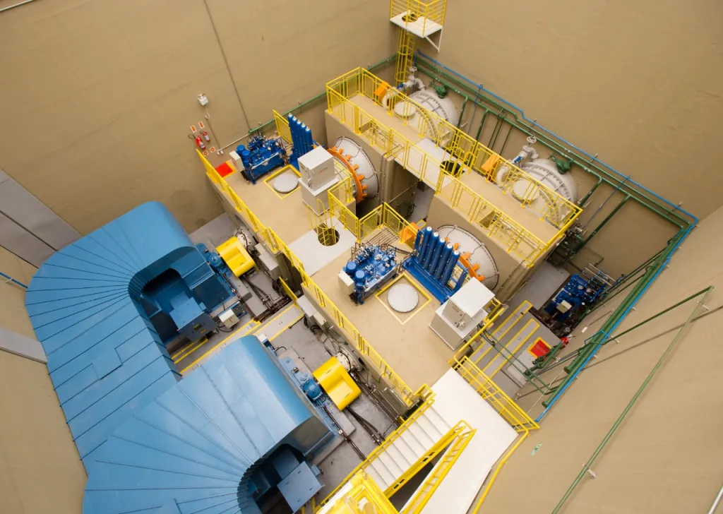 Vista aérea de uma sala industrial com máquinas grandes azuladas, tubulações verdes e estruturas metálicas amarelas.