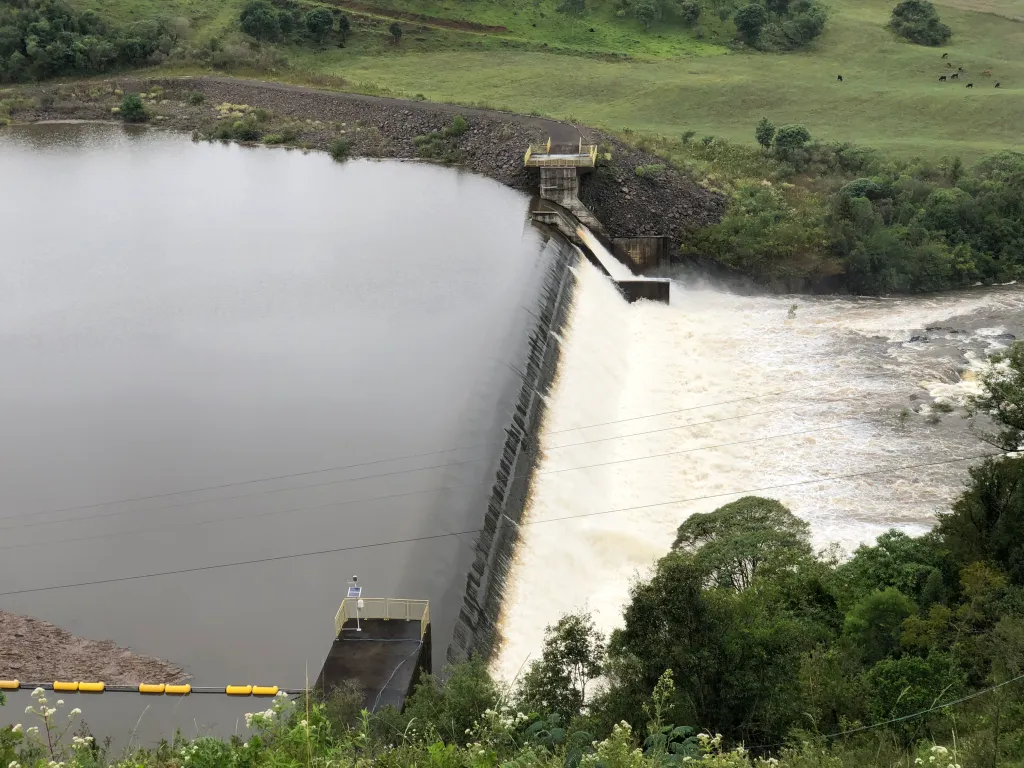 Barragem liberando água por um vertedouro com área verde ao redor.
