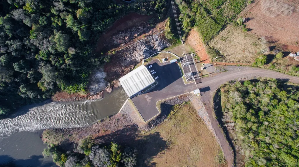 Vista aérea de uma pequena usina hidrelétrica rodeada por vegetação e campos, com um canal de água fluindo ao lado.