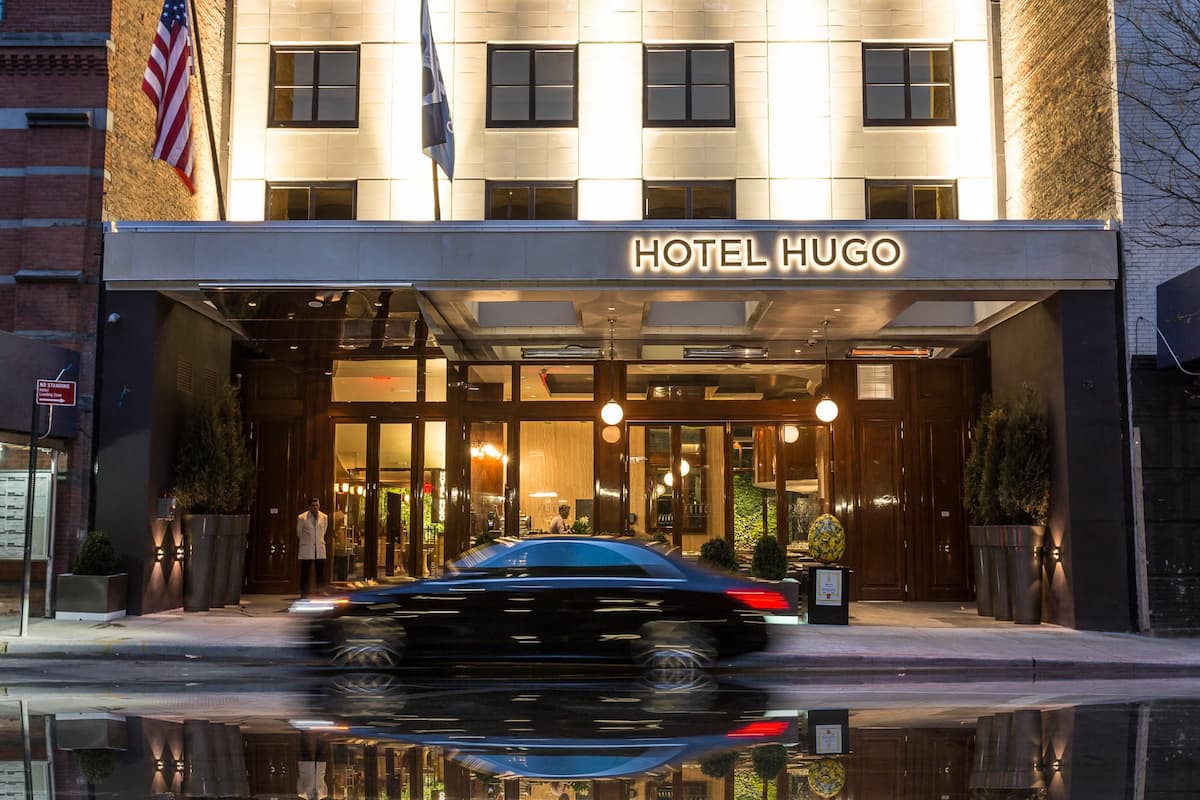 Hotel Hugo entrance at night with a black luxury car passing by, showcasing elegant lighting and flags