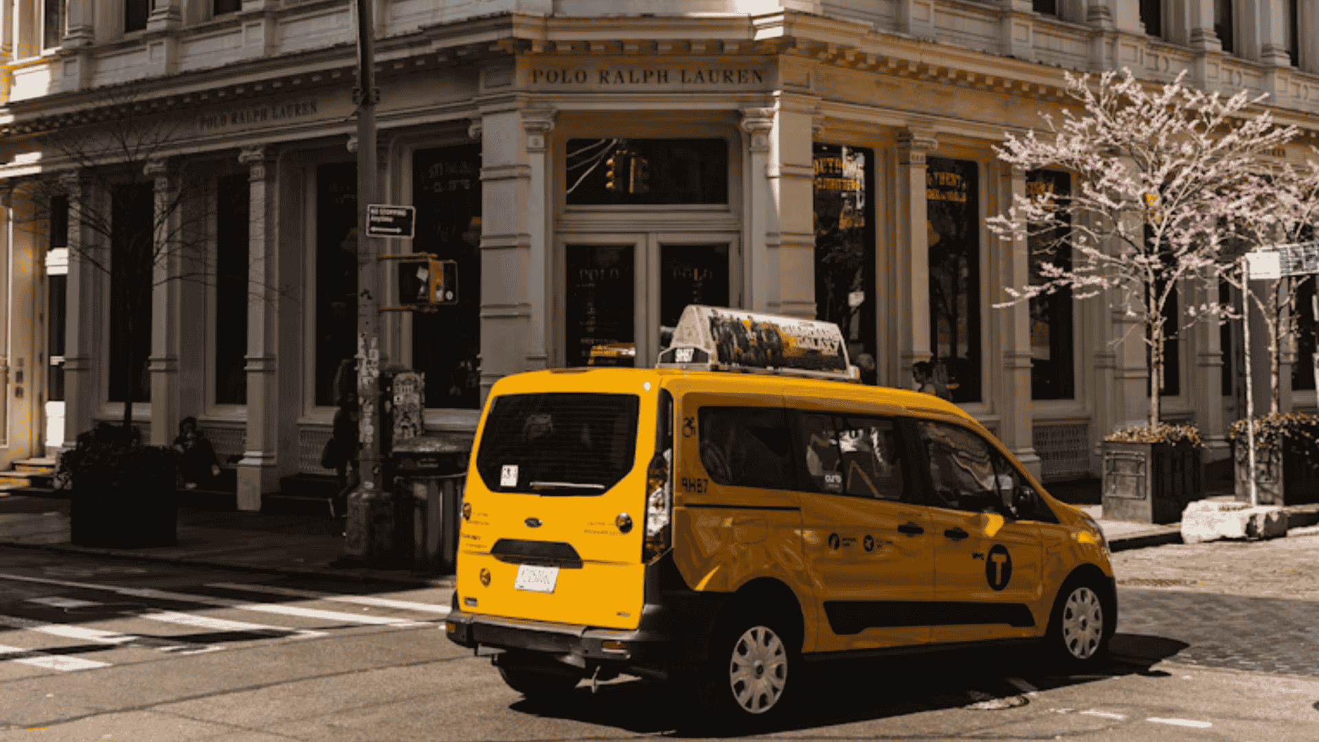 A yellow New York City taxi minivan turning at a street corner with a Polo Ralph Lauren store in the background.