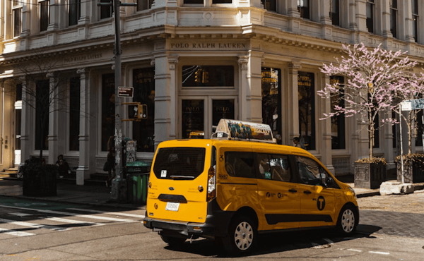 A yellow New York City taxi minivan turning at a street corner with a Polo Ralph Lauren store in the background.