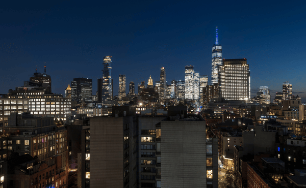 A nighttime panoramic view of the New York City skyline, featuring illuminated skyscrapers including the One World Trade Center against a dark blue sky.