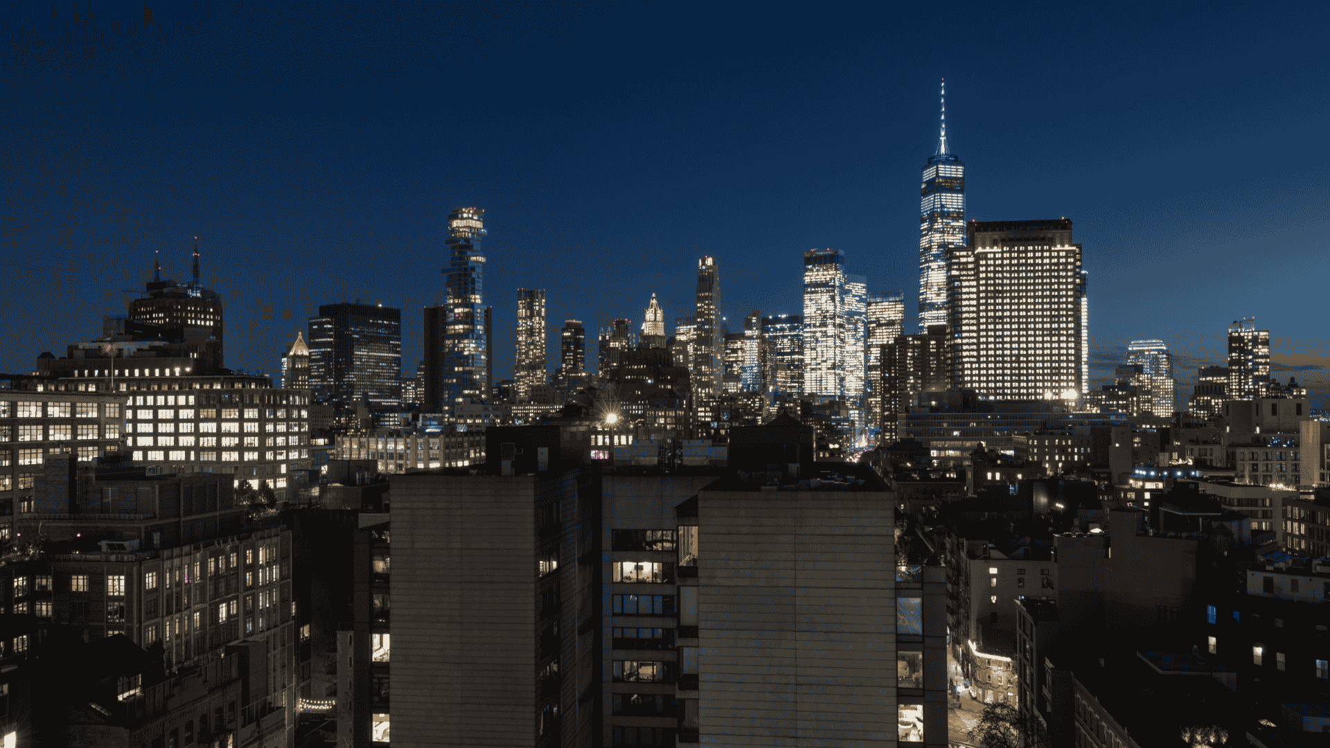 A nighttime panoramic view of the New York City skyline, featuring illuminated skyscrapers including the One World Trade Center against a dark blue sky.