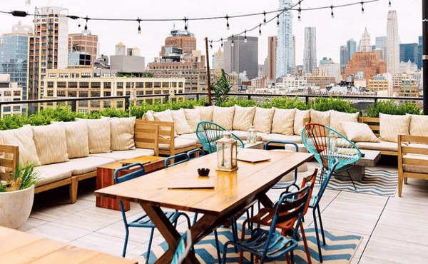 A rooftop lounge area featuring a wooden dining table, colorful metal chairs, and plush sofa seating with a view of a city skyline.
